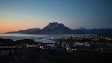 A twilight scene of Nuuk, Greenland, with houses lit up against a backdrop of mountains, featuring a mix of colourful buildings set along the coast. The sky is tinged with soft pink and orange hues as night approaches.